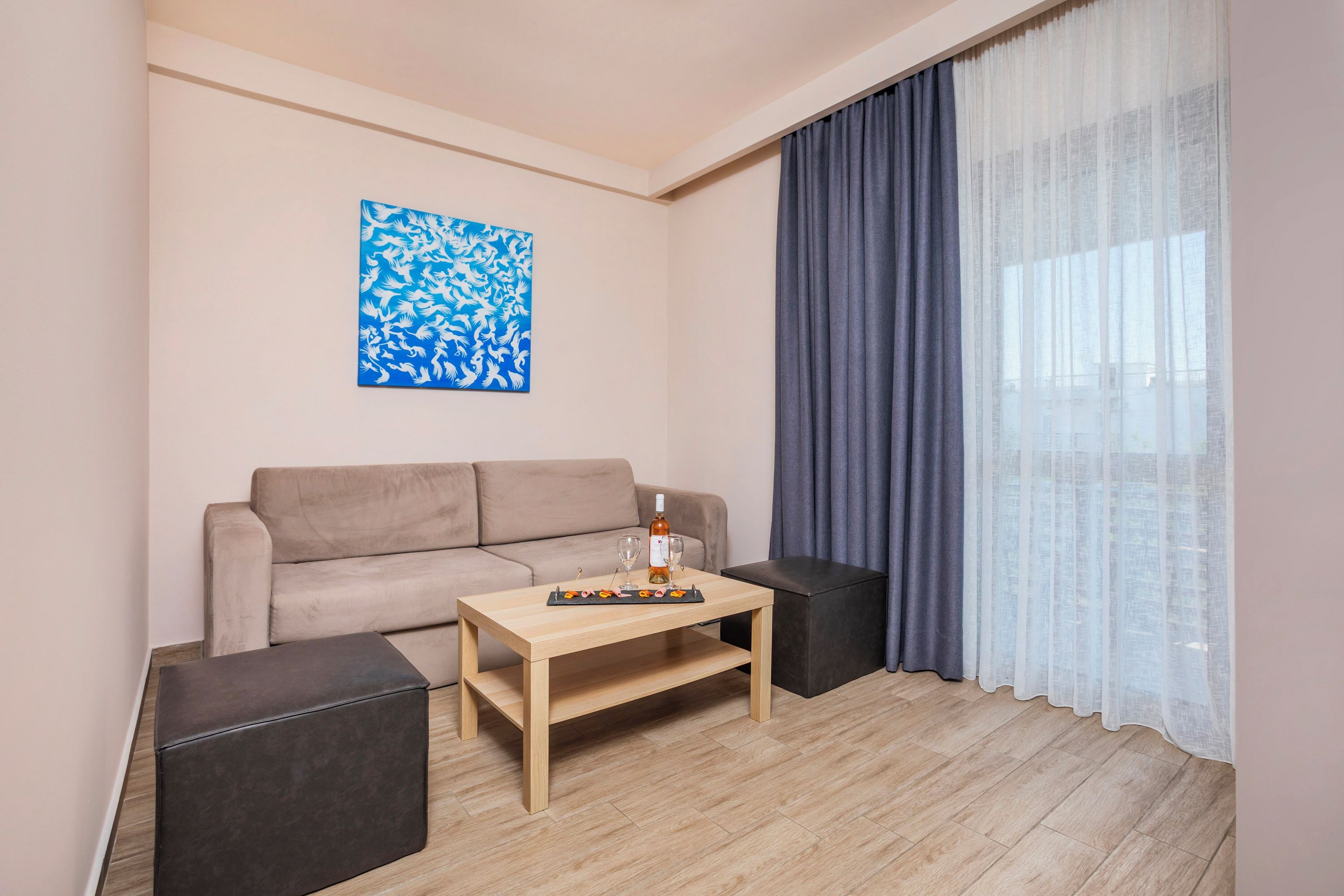 A modern living room with a beige sofa, a wooden coffee table, and a black ottoman. A blue abstract painting is on the wall, and a window with sheer curtains is visible.