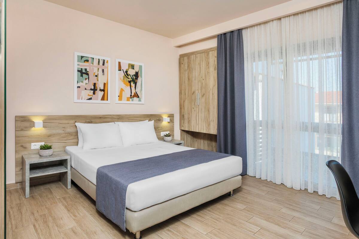 A modern hotel room with a large bed, a wooden headboard, two bedside tables with lamps, a small plant, and abstract art on the wall.