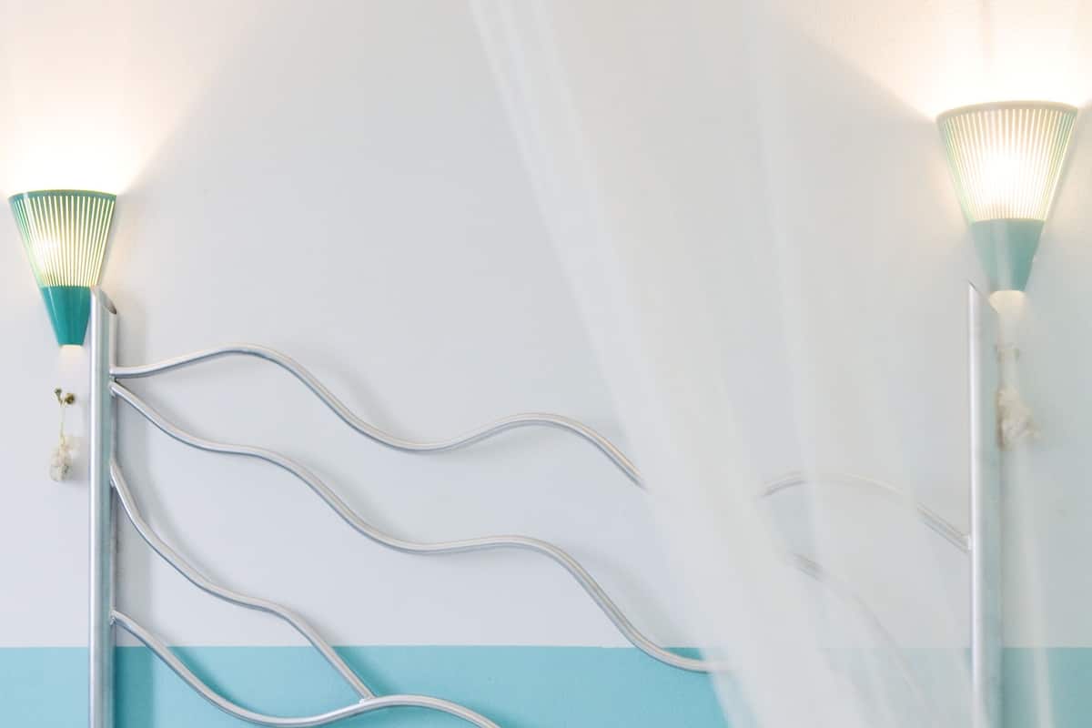 A bed with white bedding, a metal headboard with wavy lines, and two wall-mounted lamps with green shades.