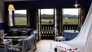 A bedroom with a large bed, a sitting area with a bench and chairs, a small table with a vase of flowers, and a view of a green field through open doors. - Hotel Rural La Llosa de Fombona (Gozon)