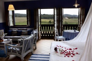 Quarto com cama grande, área de estar com banco e cadeiras, pequena mesa com vaso de flores e vista para um campo verde através de portas abertas.
