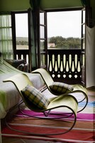 A room with a bed, two rocking chairs, a colorful striped rug, and a balcony with a view of trees and a house.