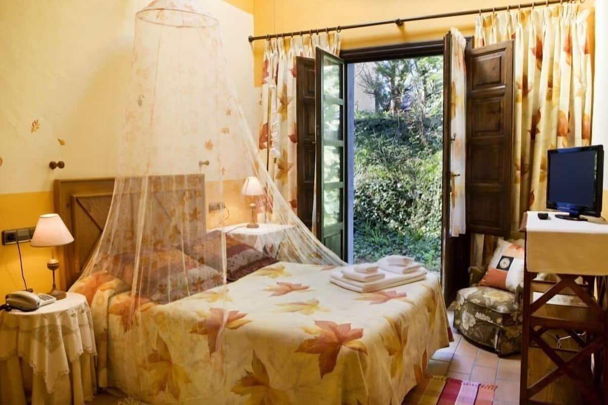 A bedroom with a bed, a bedside table with a lamp, a TV, and a window with curtains.