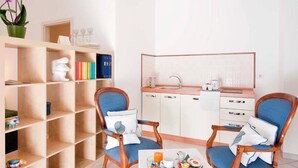 A compact room with a kitchenette, two blue upholstered chairs, a small table with breakfast items, and a bookshelf with various items. - A Due Passi (Forte dei Marmi)