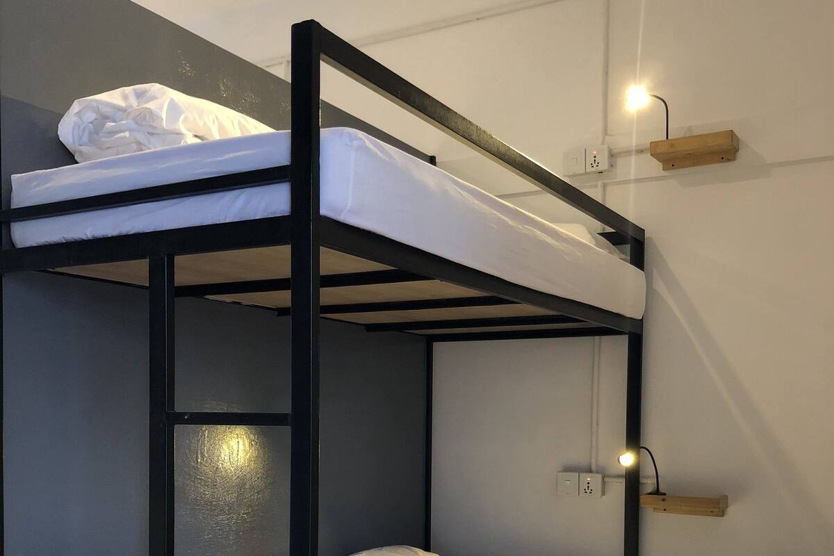 A bunk bed with white bedding, a wall socket, and a wall-mounted light fixture.