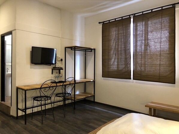 A compact room with a bed, a desk with two chairs, a TV mounted on the wall, and a window with brown curtains.