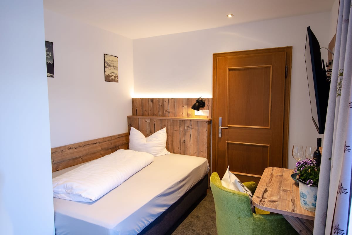 Une petite chambre à coucher, bien rangée, avec un lit simple, une tête de lit en bois, une table de chevet avec une lampe, une chaise verte et une petite table sur laquelle est posée une plante en pot.
