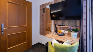 A compact room with a wooden door, a flat-screen TV mounted on the wall, a small wooden table with a bowl of fruit, a bottle of wine, and a potted plant. - Gasthof Messerschmied (Grassau)