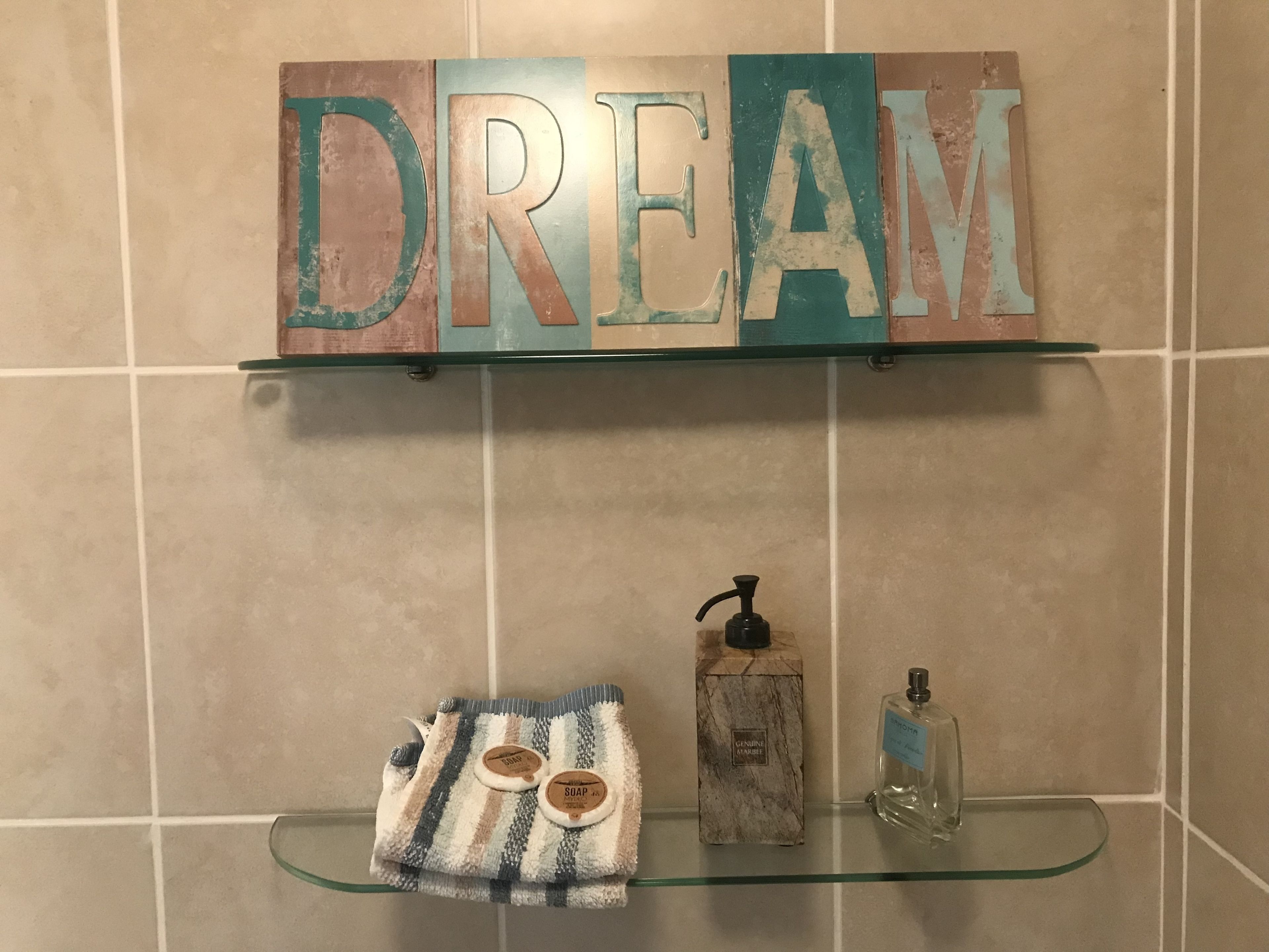 A bathroom with a shelf displaying the word "DREAM" in large letters, a striped towel, a soap dispenser, and a bottle of liquid soap.