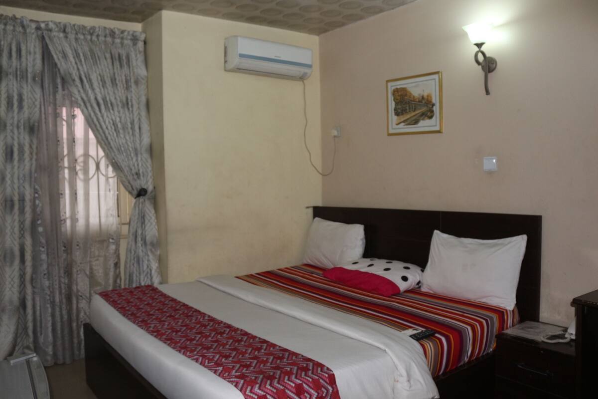 Una habitación de hotel con cama, mesita de noche, equipo de aire acondicionado, luz en la pared y ventana con cortinas.