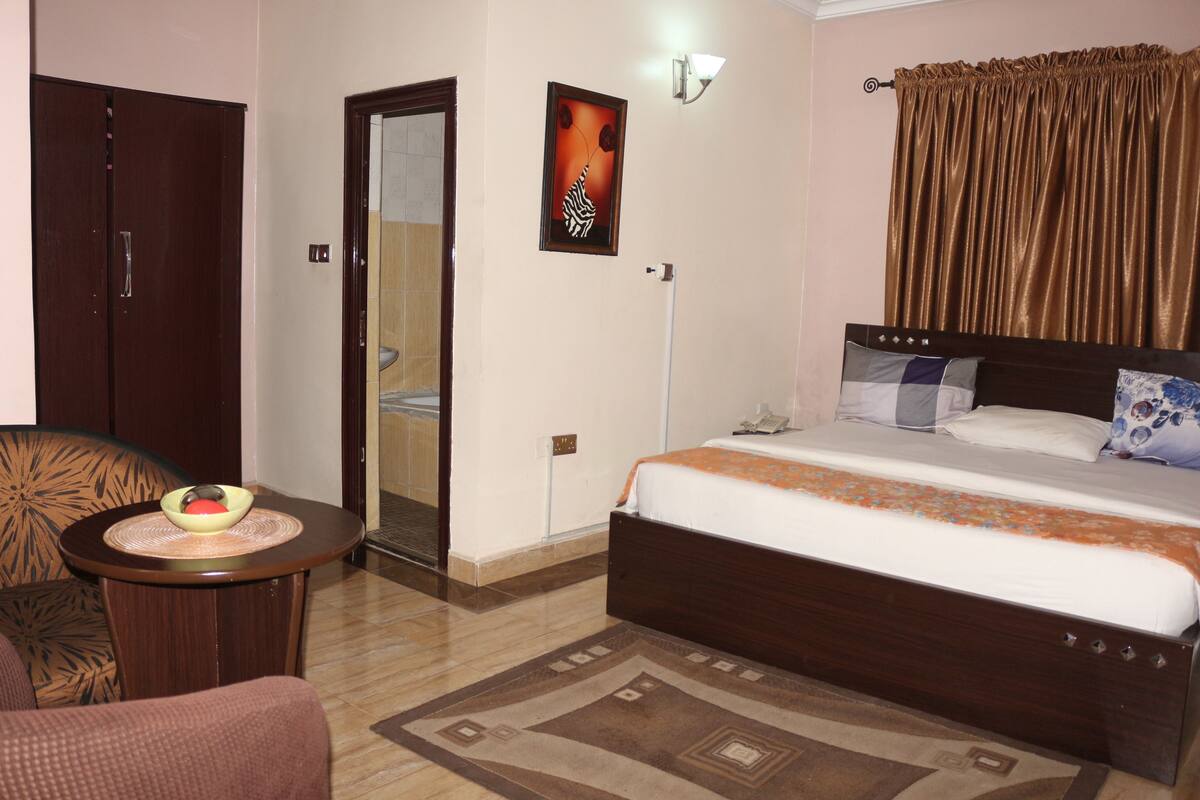 Una habitación de hotel con una cama, una silla, una mesita con un bol, una lámpara de pared y una puerta.