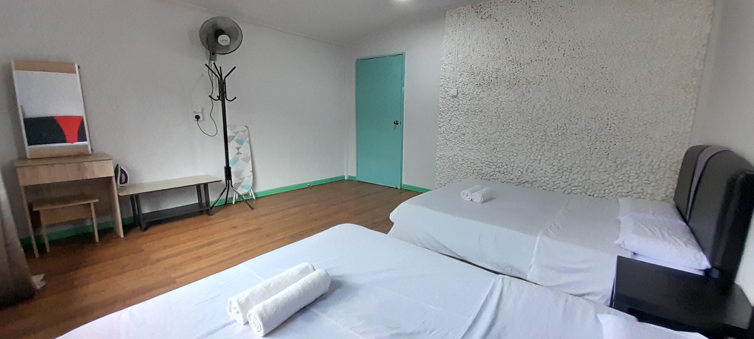A hotel room with two beds, a desk, a chair, a fan, and a door.