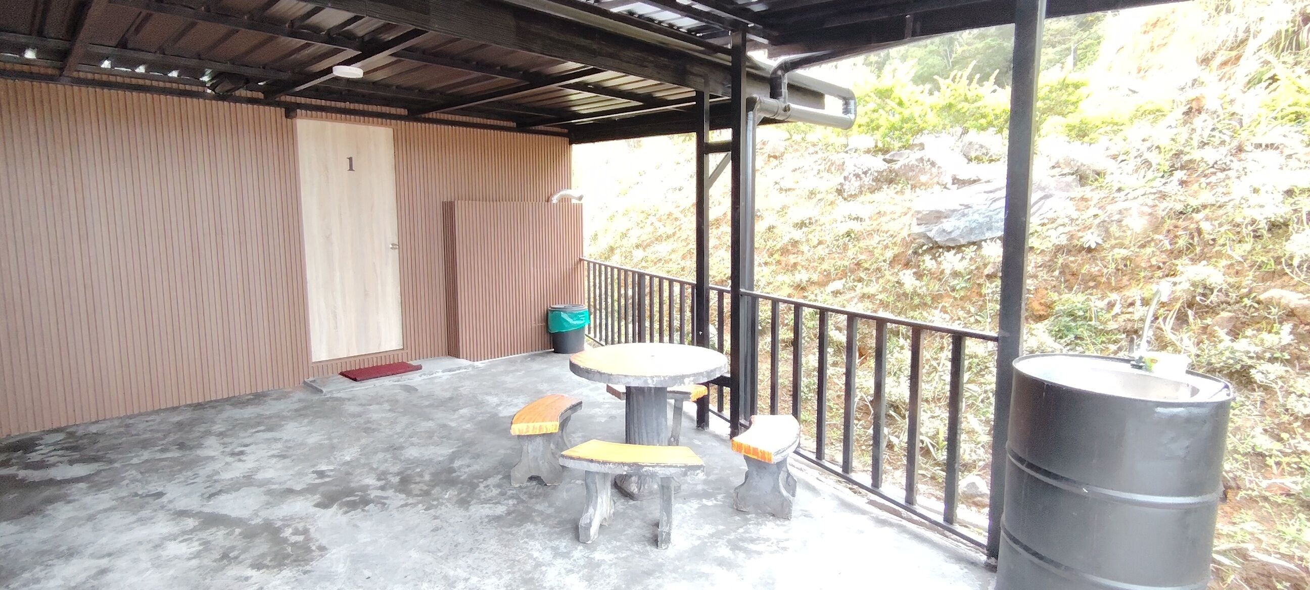 Un espacio al aire libre techado con piso de concreto, una mesa redonda con bancos, un basurero y una baranda metálica.