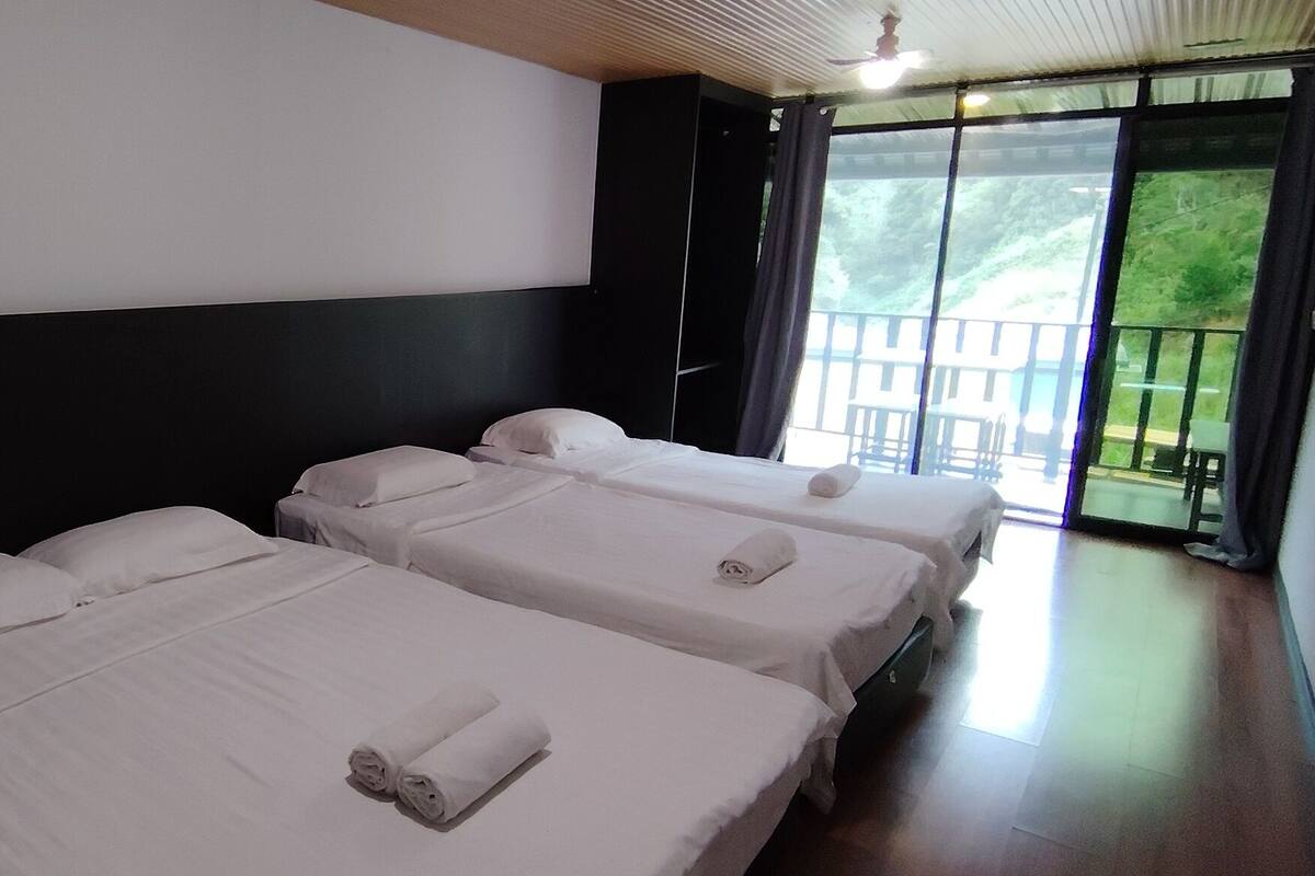 A hotel room with three beds, a small table with red cups, a white chair, and a large window with a view of greenery.
