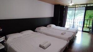 Habitación de hotel con tres camas, una mesita con vasos rojos, una silla blanca y un ventanal con vistas a zonas verdes.