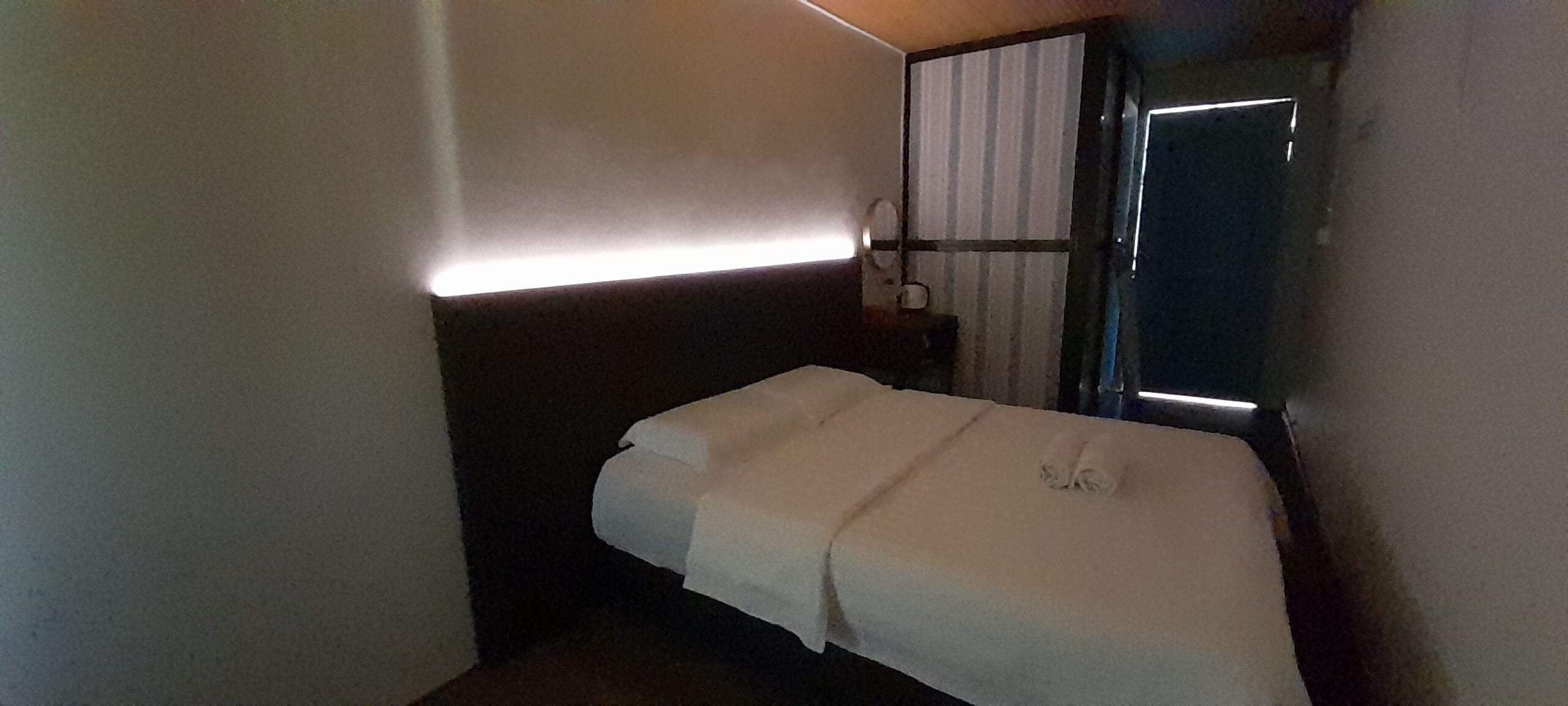 A small, dimly lit hotel room with a single bed, a nightstand, and a window with curtains.