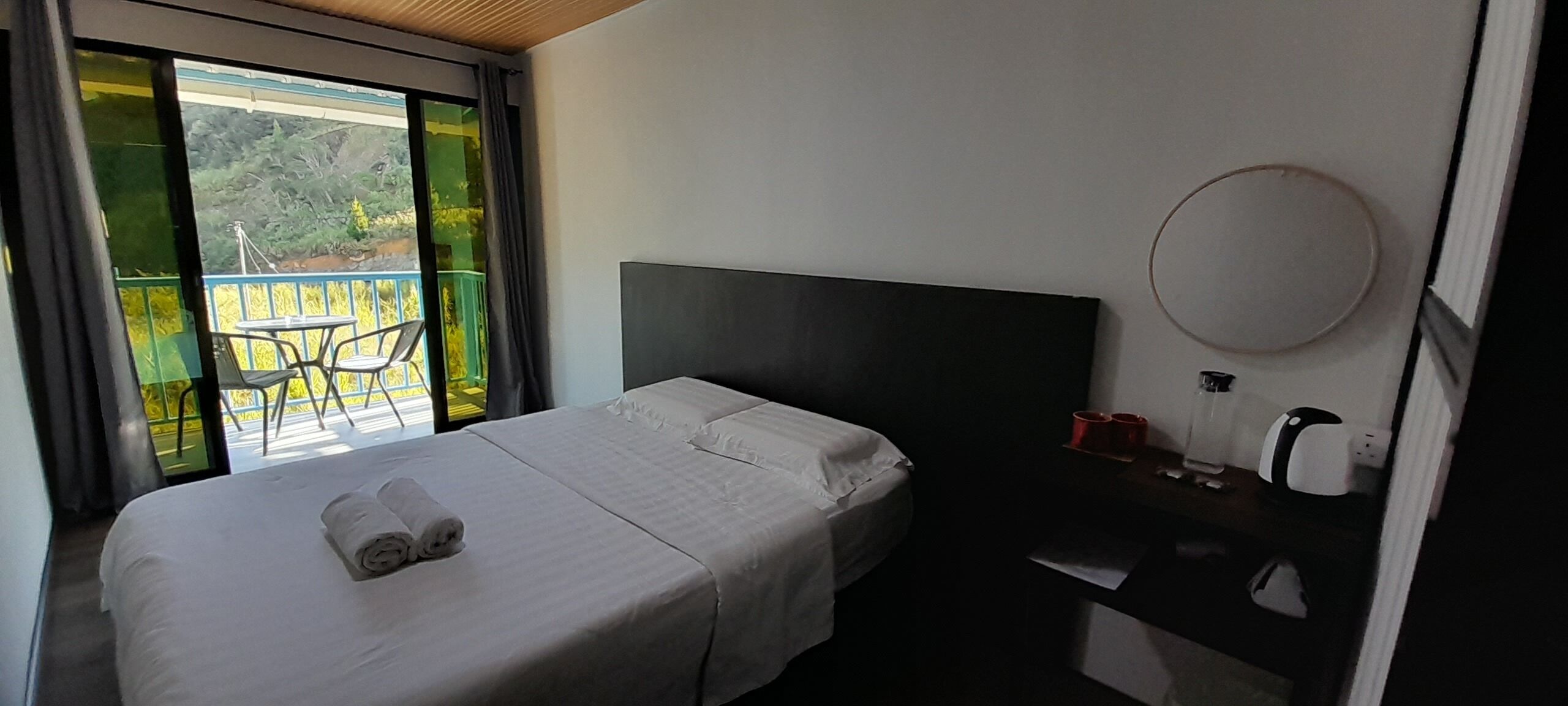 A hotel room with a bed, a nightstand, a round mirror, and a view of a balcony with chairs and greenery.