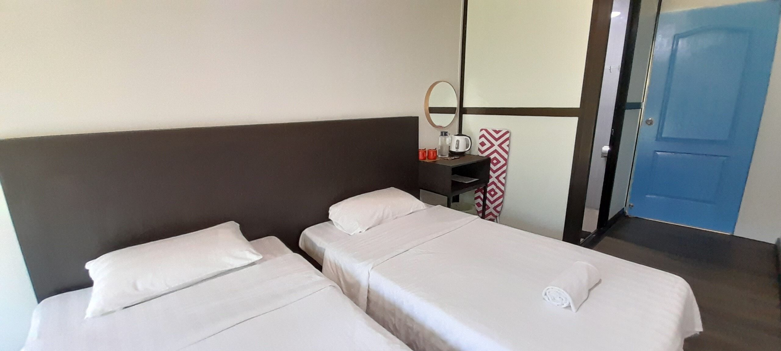 Ein Hotelzimmer mit zwei Einzelbetten, einem Nachttisch mit Spiegel, einem rot-weißen Wandbehang und einer blauen Tür.