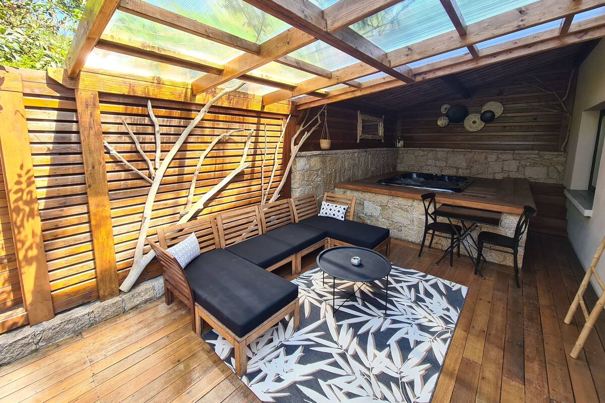 A wooden deck with a glass ceiling, a stone wall, a hot tub, and a dining area with wooden furniture.