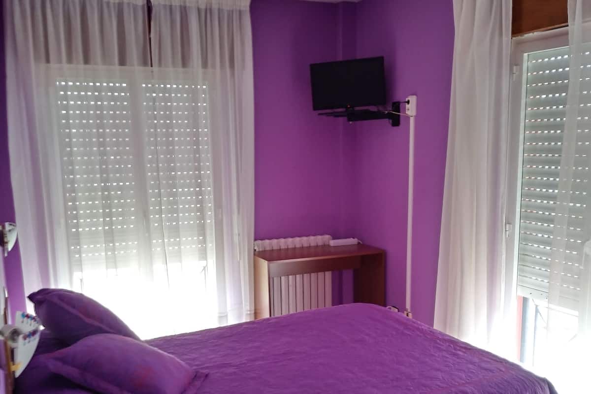 Ein Zimmer mit einem violetten Bett, einem an der Wand befestigten Fernseher und einem Fenster mit Jalousien.