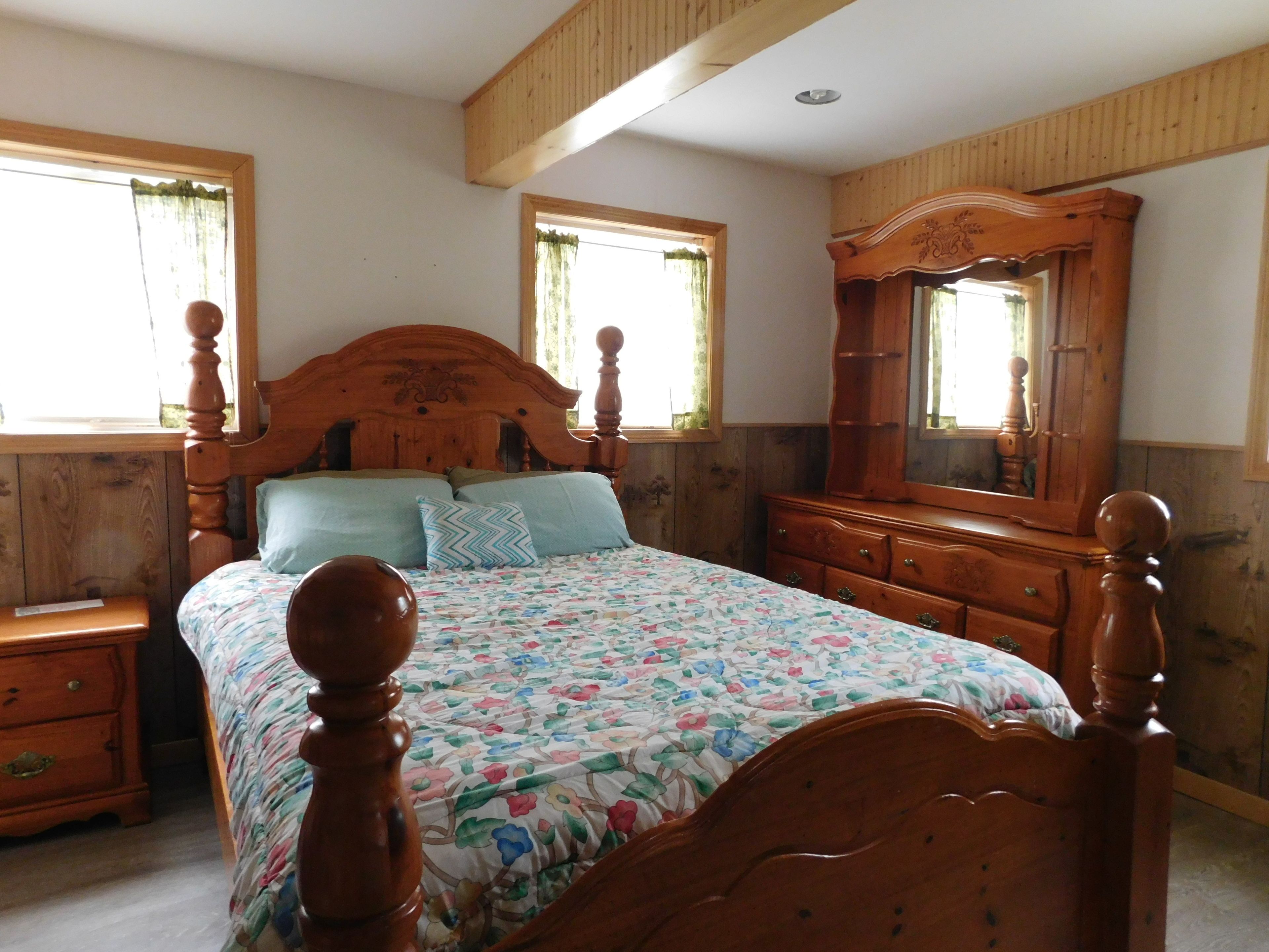 Quarto com cama de madeira, cômoda e espelho. Possui duas janelas com cortinas.