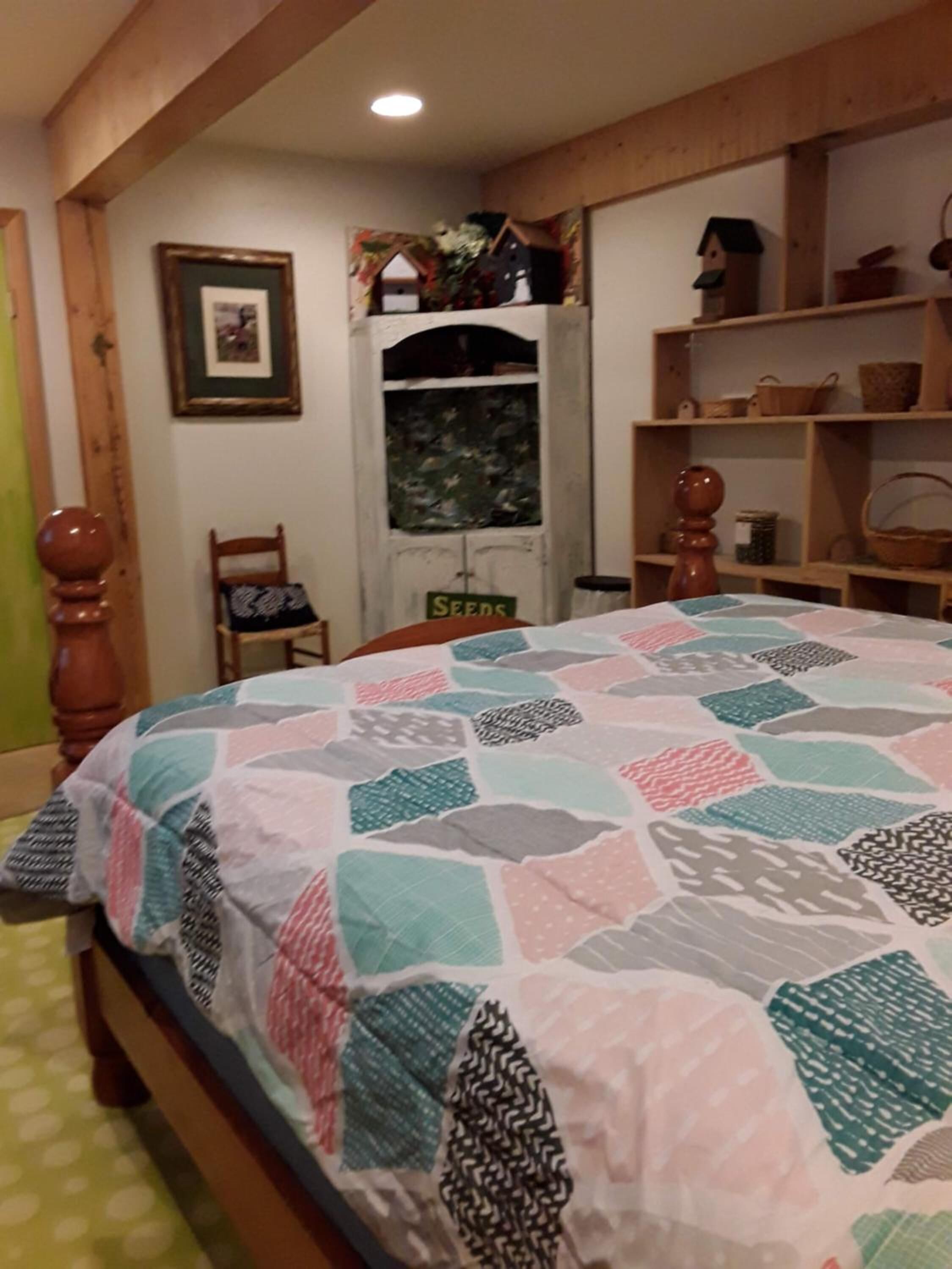 A bedroom with a bed, a chair, a shelf with baskets, a framed picture, and a window with a 'Seeds' sign.