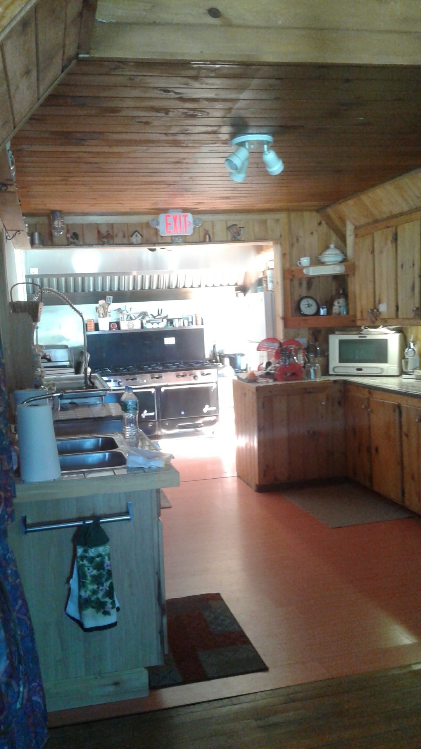 Een rustieke keuken met houten wanden en een houten plafond. Er staat een fornuis, een gootsteen en een magnetron op het aanrecht. Een vluchtroutebord is zichtbaar aan de muur.