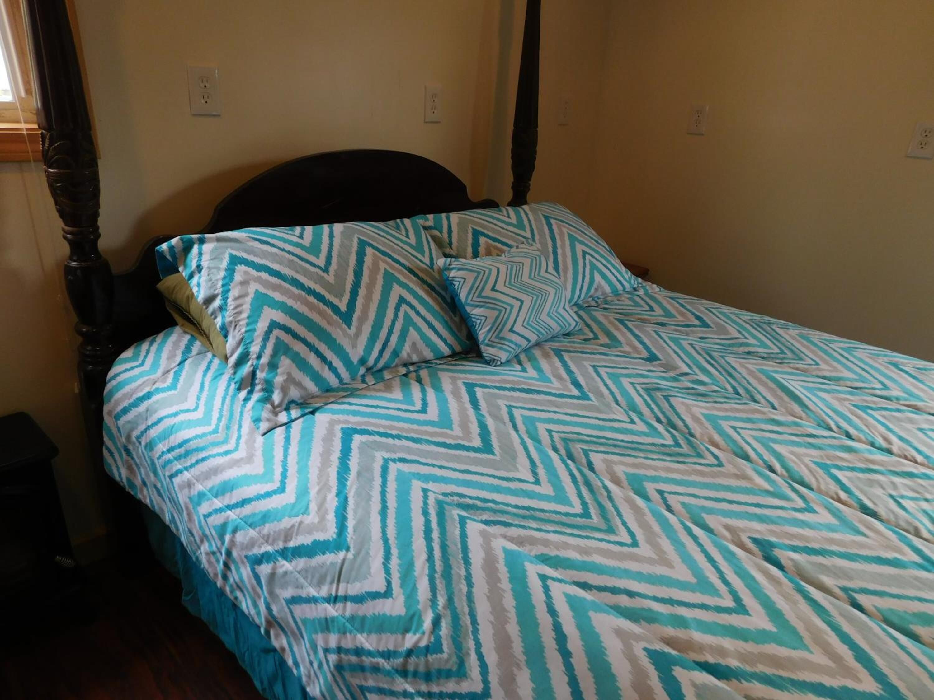 A four-poster bed with a chevron-patterned bedspread and pillows, a wooden headboard, and a plain wall with electrical outlets.