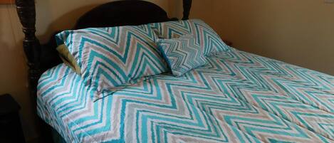 A four-poster bed with a chevron-patterned bedspread and pillows, a wooden headboard, and a plain wall with electrical outlets.