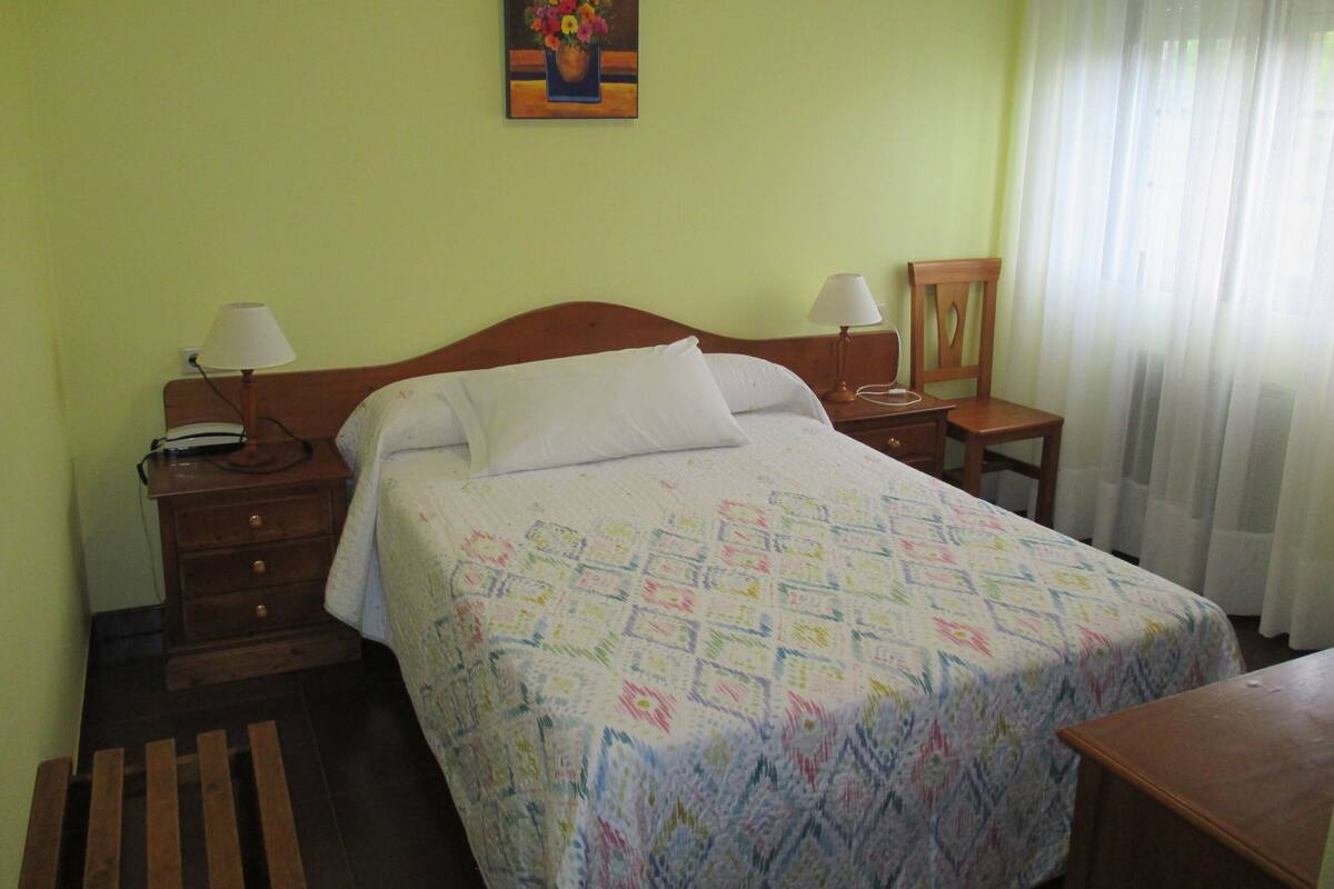 Habitación de hotel con cama, dos mesitas de noche con lámparas, un tocador de madera, una silla y un cuadro en la pared.