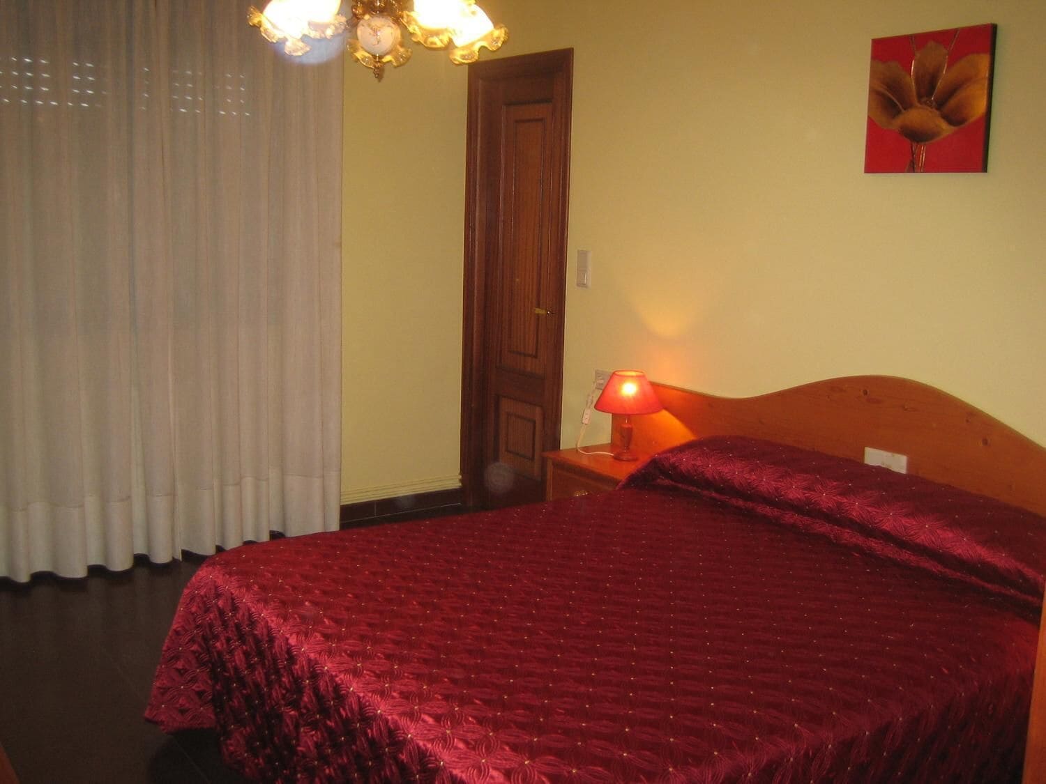 Ein Hotelzimmer mit einem großen Bett, einem hölzernen Kopfteil, einem Nachttisch mit Lampe, einer Tür und einem Fenster mit Vorhängen.