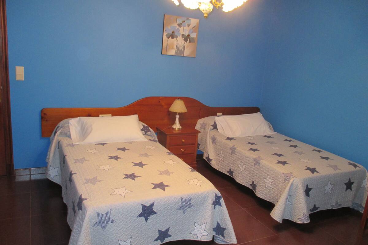 Dos camas individuales con ropa de cama estampada con motivos de estrellas, un cabecero de madera, una mesita de noche con lámpara y un cuadro colgado en la pared.