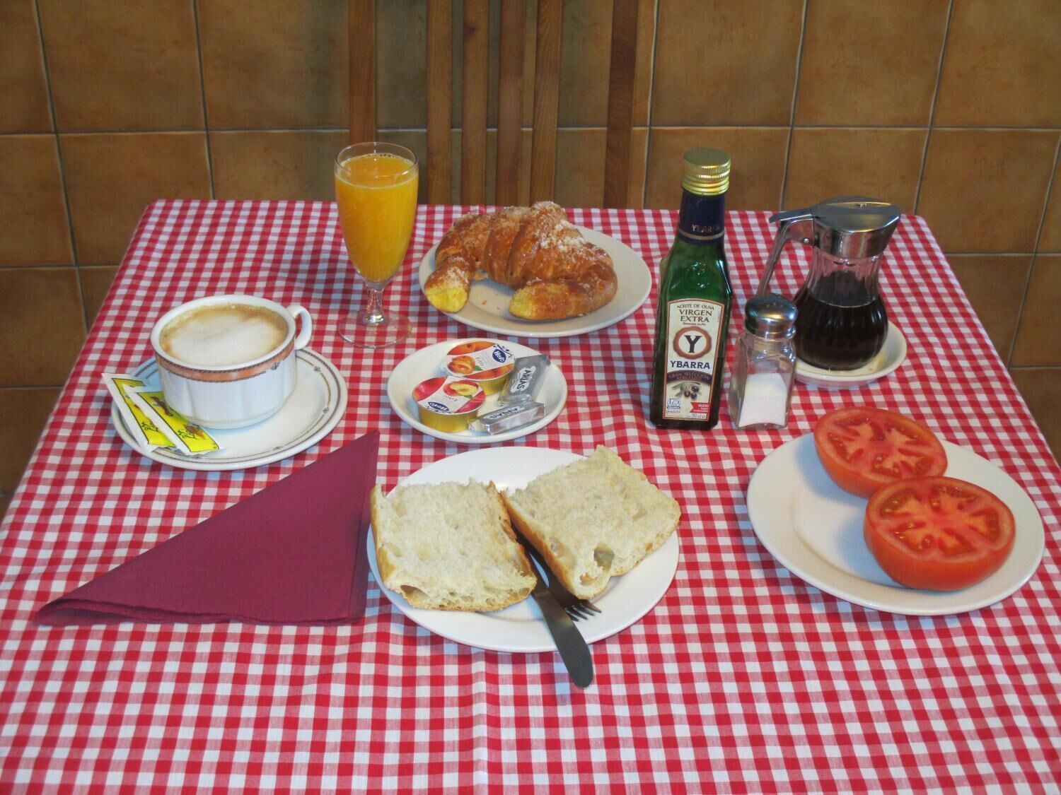 A table with a red and white checkered tablecloth, a croissant, a glass of orange juice, a bottle of Ybarra sauce, a pitcher of black coffee, a plate with bread and tomato slices, and a cup of coffee.