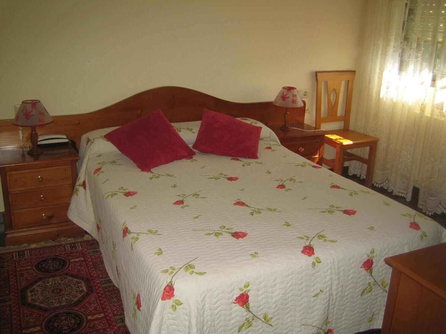 A neatly made bed with a floral bedspread and two red pillows, a wooden nightstand with a lamp, a wooden chair, and a small table with a lamp.