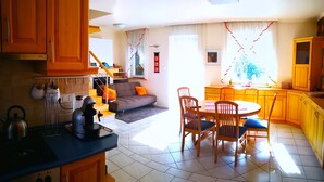 A kitchen with a stove, kettle, and coffee maker. There is a dining area with a table and chairs. A sofa is in the background. - Aurora Premium Home (Gyor)
