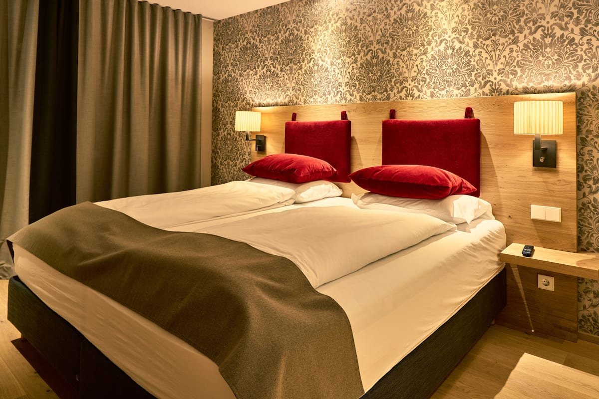 A hotel room with a large bed, red pillows, a wooden headboard, and a nightstand with a lamp.