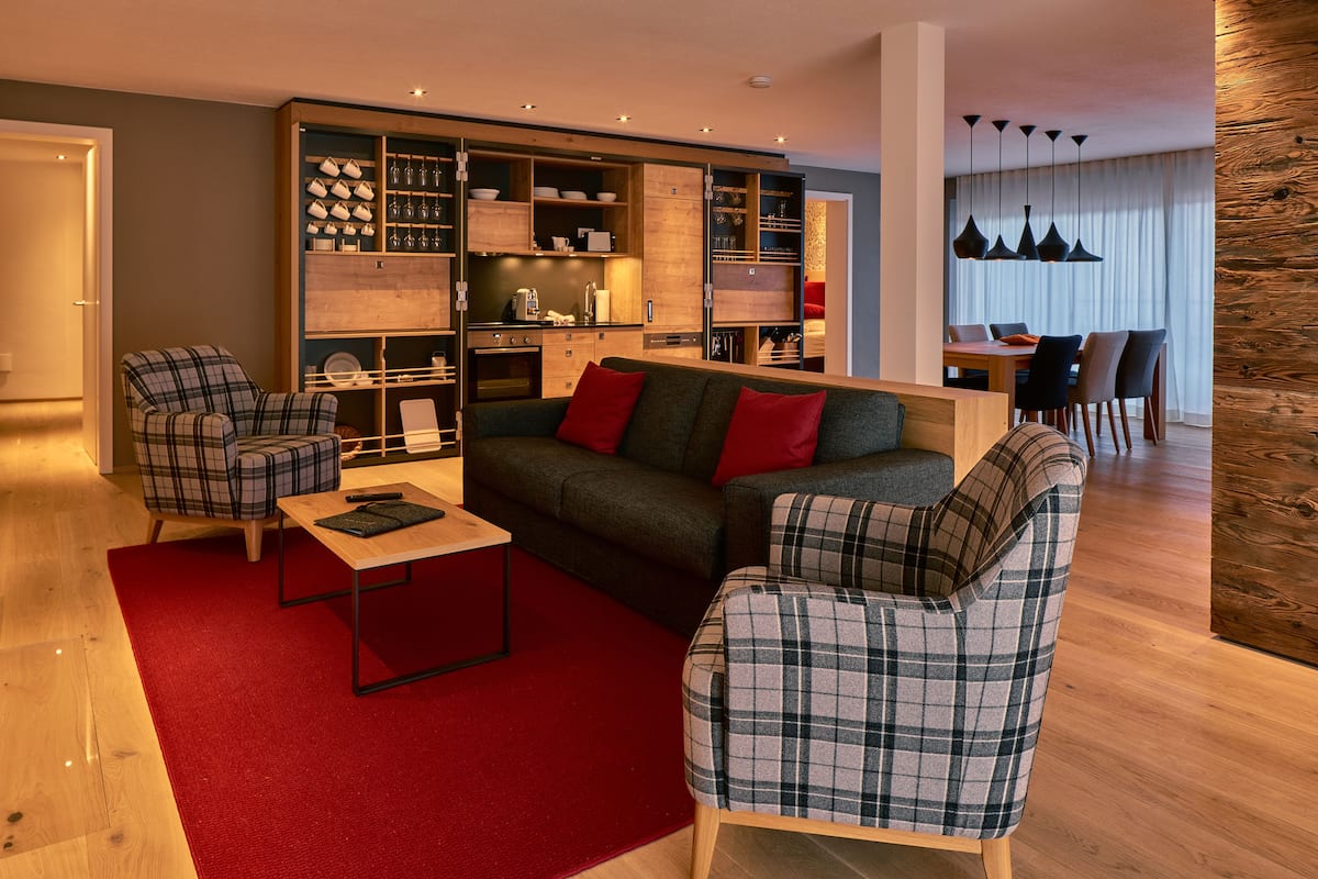 A modern living room with a sofa, two plaid armchairs, a coffee table, and a built-in wooden shelving unit with kitchenware.
