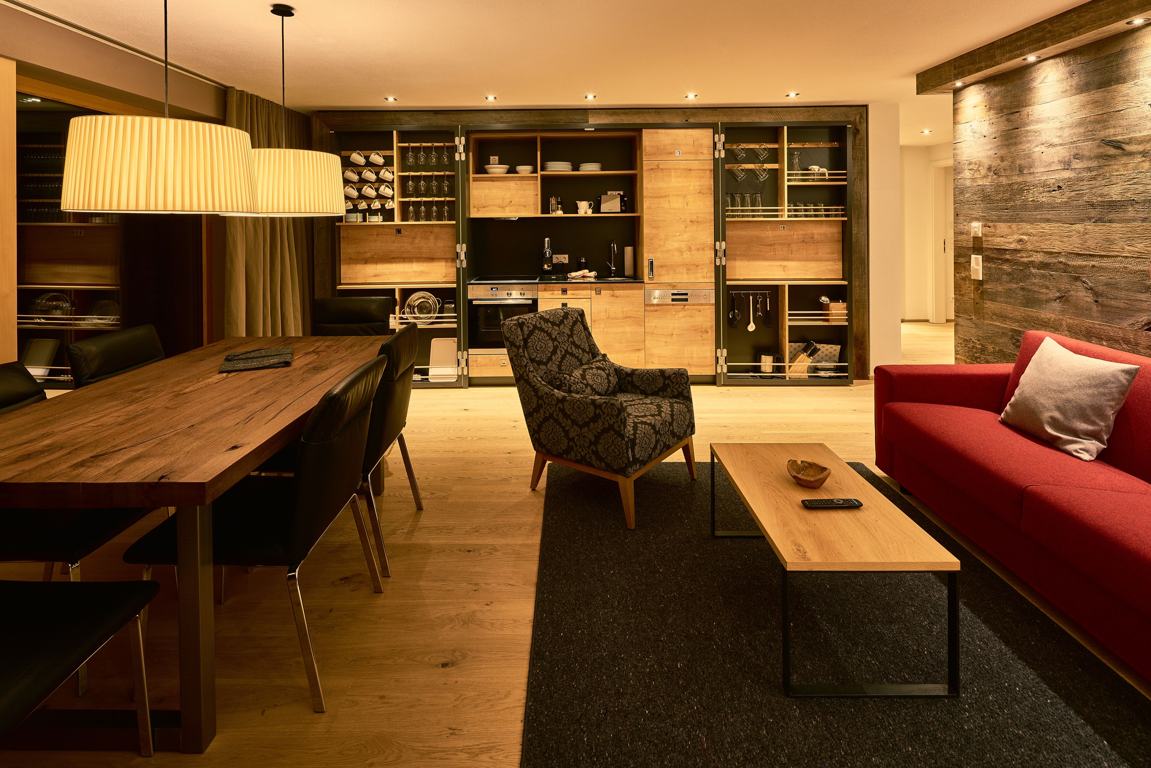 Una sala de estar moderna con una mesa de comedor de madera, un sofá rojo, un sillón estampado y una zona de cocina con armarios y estanterías de madera.