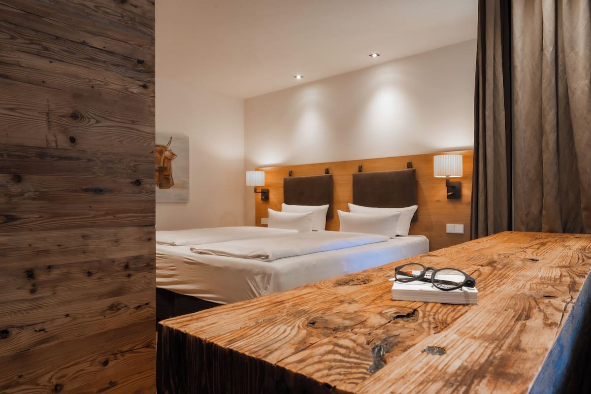 A hotel room with a wooden headboard, two beds with white linens, a wooden table with glasses and a book, and a wall-mounted lamp.