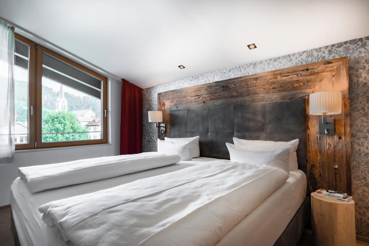 A hotel room with two beds, a large window with a view of a church and greenery, and a wooden headboard.