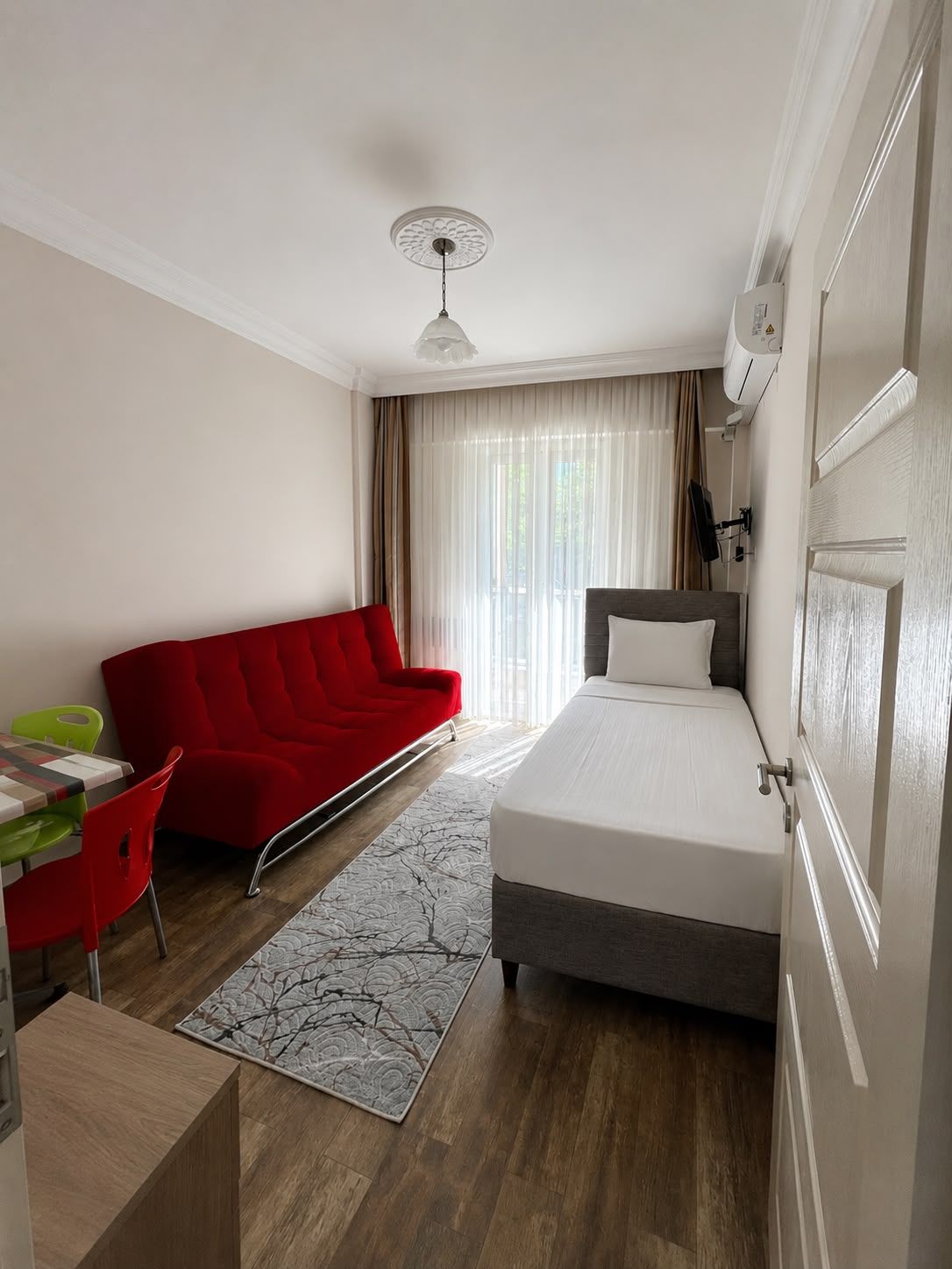 A hotel room with a bed, a red sofa, a dining table with chairs, and a window with curtains.