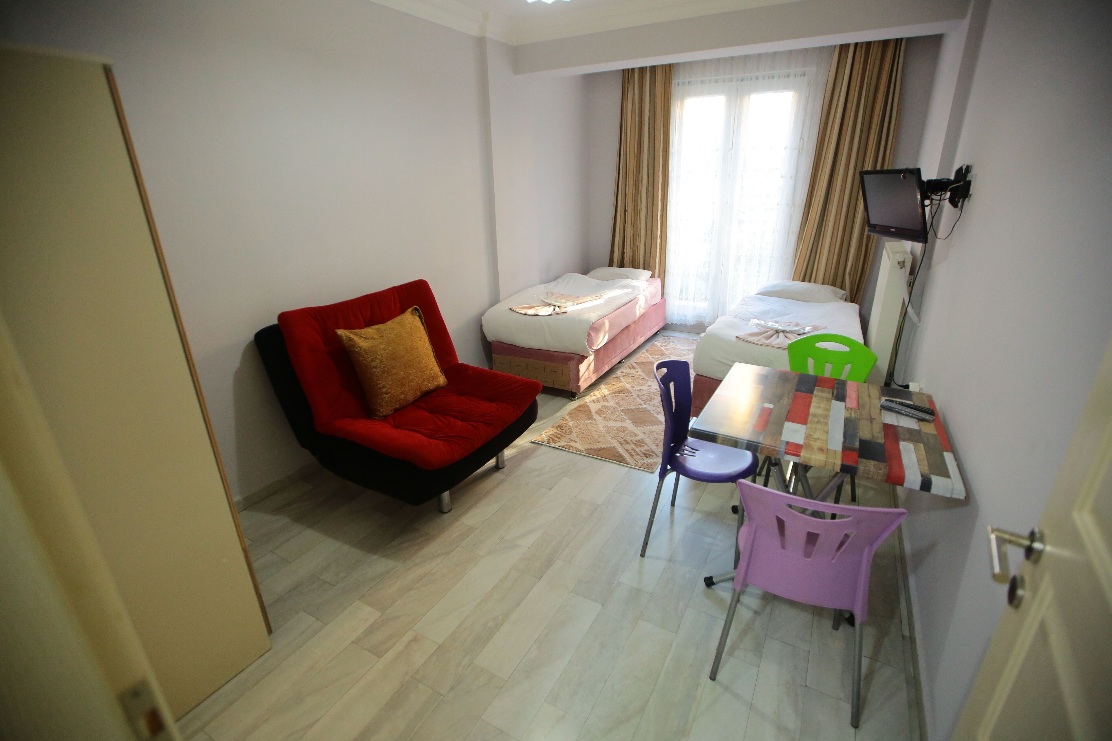 A small, well-lit room with a red chair, a bed, a desk with a chair, and a TV mounted on the wall.