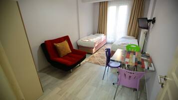 A small, well-lit room with a red chair, a bed, a desk with a chair, and a TV mounted on the wall.