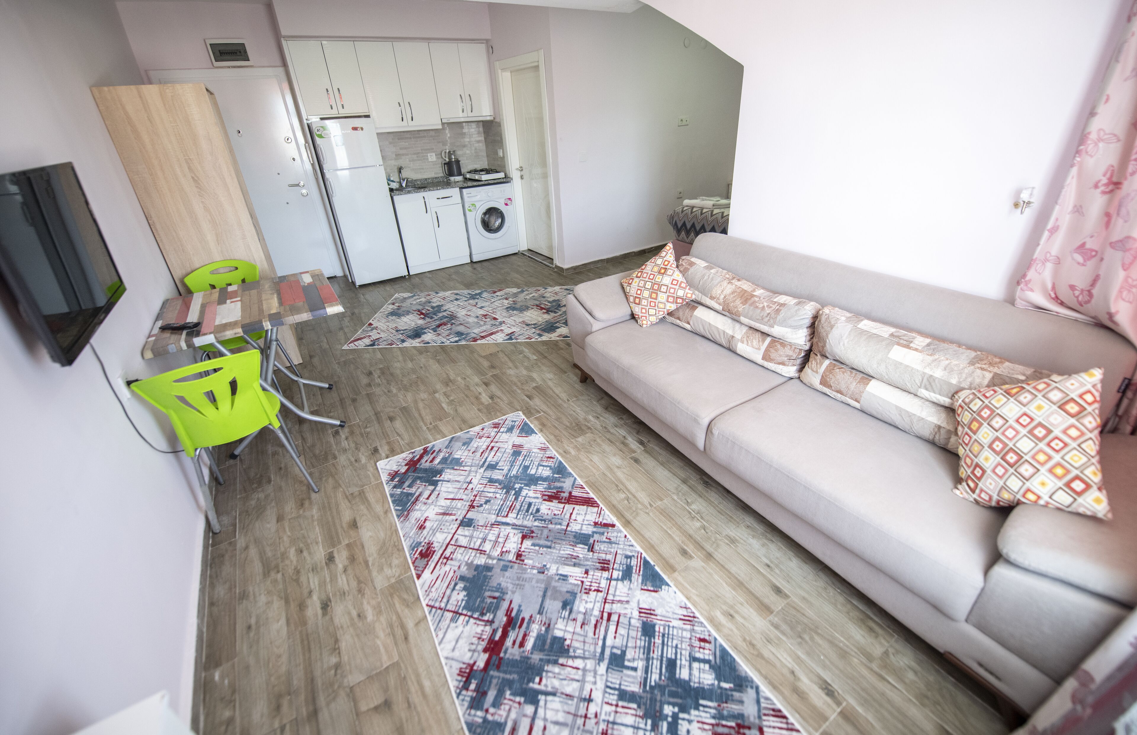 A compact living space with a sofa, a dining table, a green chair, a TV, a kitchen area, and a patterned rug.