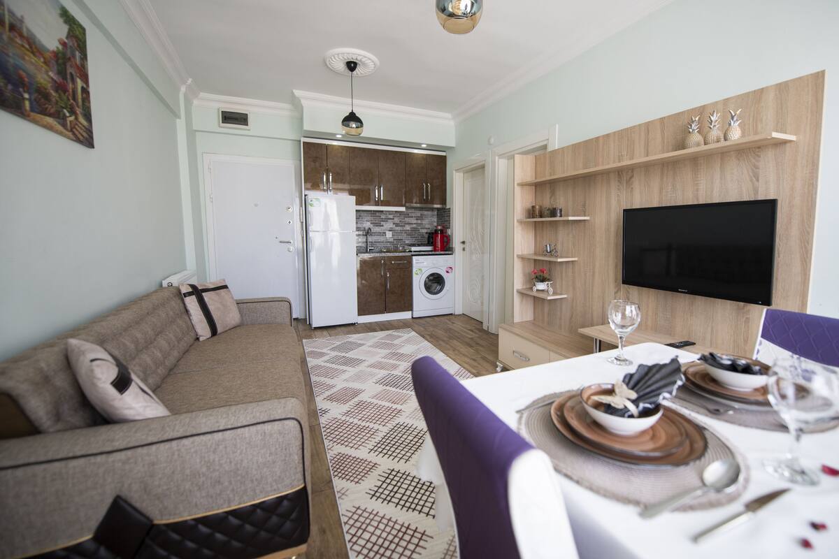 A modern living room with a sofa, a dining table set for two, and a kitchen in the background.