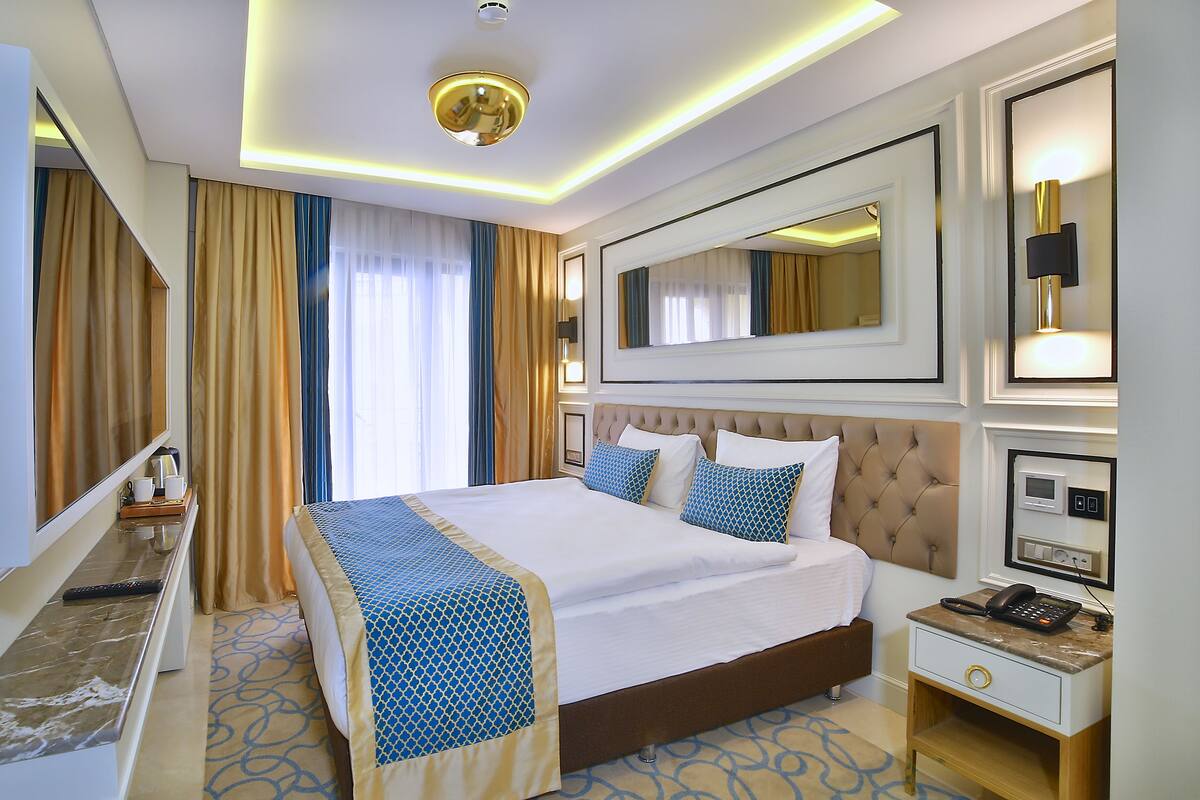 Una habitación de hotel con una cama grande, una mesita de noche con un teléfono y una lámpara, un espejo y una ventana con cortinas.