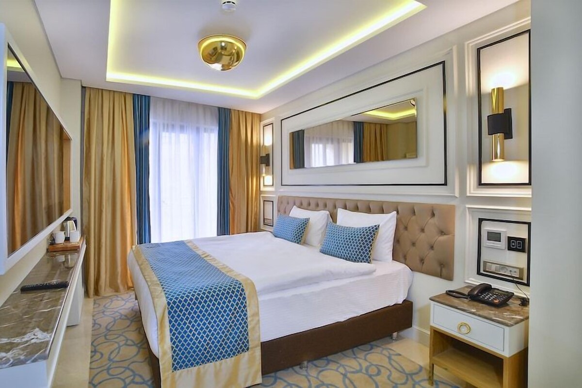 A hotel room with a large bed, a bedside table with a phone, a mirror, and a window with curtains.