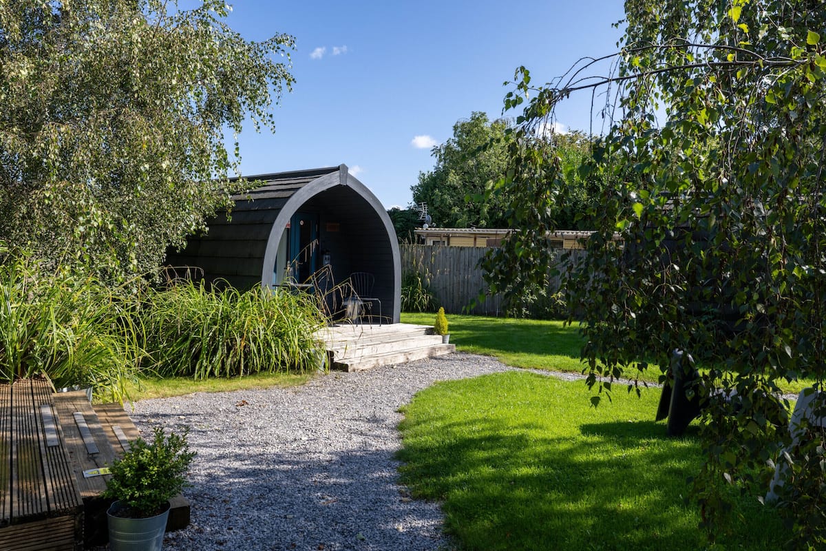 Un chalet rustique doté d’un toit incurvé, d’un chemin de gravier menant à l’entrée et d’une terrasse en bois agrémentée de plantes en pot.