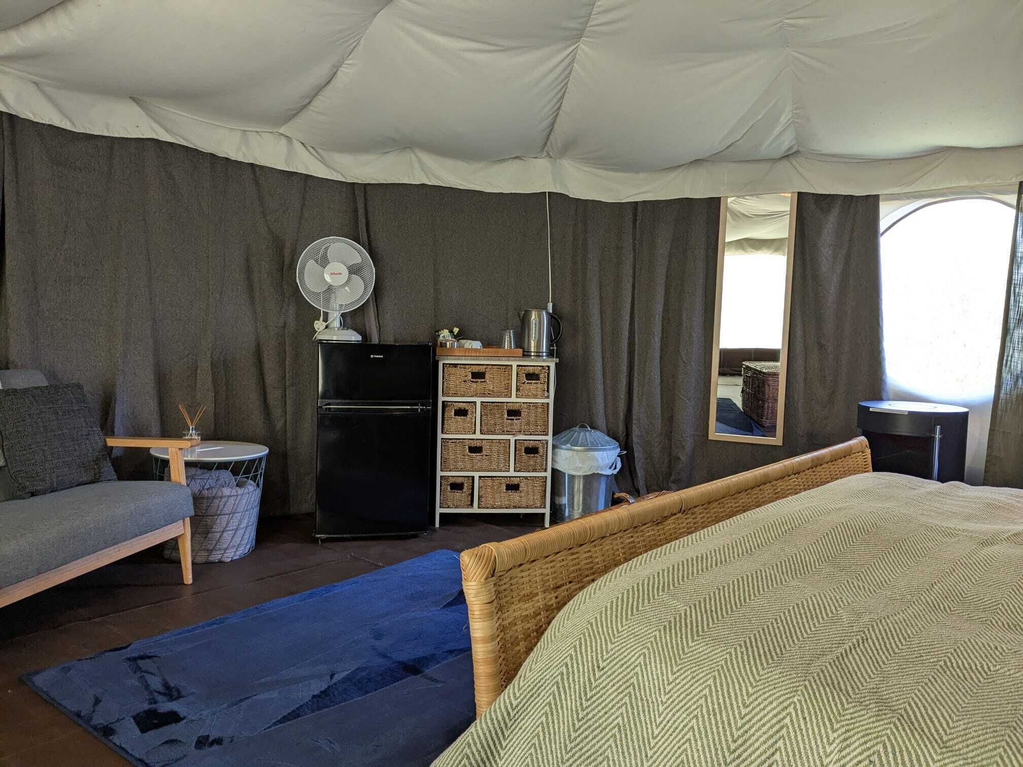 Una tenda arredata con un letto, un tavolino, una ventola, un frigorifero e cesti portaoggetti.