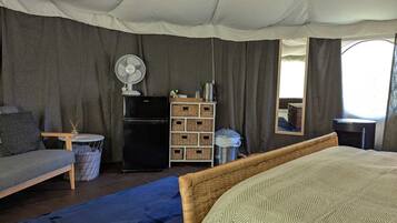 Una tenda arredata con un letto, un tavolino, una ventola, un frigorifero e cesti portaoggetti.